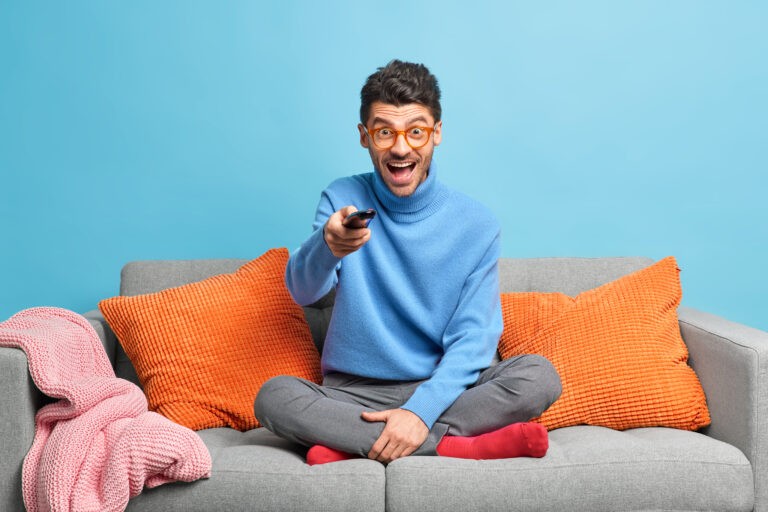 people-leisure-pastime-concept-overjoyed-unshaven-adult-man-sits-in-lotus-poses-on-sofa-holds-remote-control-and-watches-funny-show-on-television